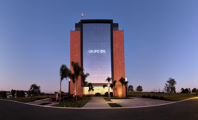 Fachada do edifício do Grupo OM ao entardecer, com iluminação destacando o logotipo “GRUPO OM” refletido no vidro central. O céu em tons de lilás e laranja contrasta com a estrutura espelhada e as palmeiras à frente, criando uma atmosfera elegante e simbólica do dinamismo e da visão de futuro das empresas que fazem parte do grupo.