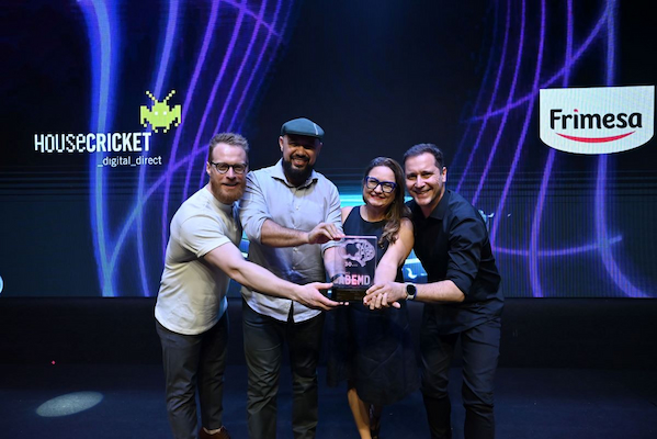 Profissionais da HouseCricket posam juntos no palco segurando o troféu ABEMD, em celebração ao case premiado desenvolvido em parceria com a Frimesa. No telão ao fundo aparecem os logotipos das duas marcas, destacando a sinergia entre agência e cliente. A foto captura um momento de orgulho e conquista, refletindo a excelência das estratégias digitais e o reconhecimento do trabalho conjunto.